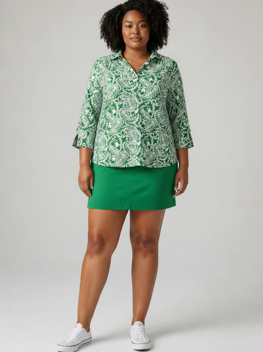 A woman modeling a green and white paisley patterned button-down shirt with three-quarter sleeves, paired with a solid green mini skirt and white sneakers, against a light gray studio background.