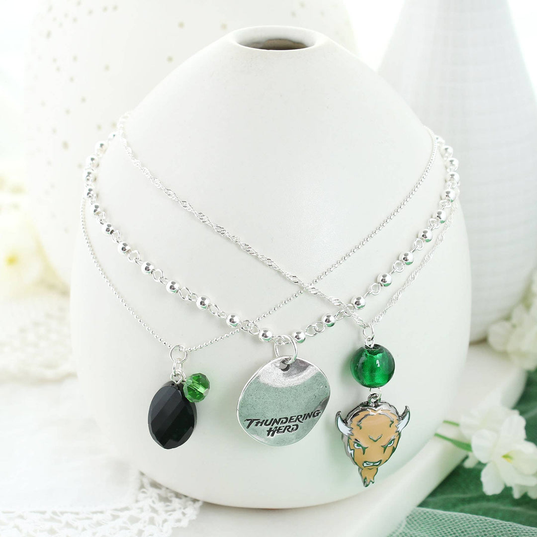 Set of three silver plated 16” necklaces (each with a claw clasp and 3” extender). Each necklace features a different chain style and charm, to include green and black beads, an enamel Marshall logo charm, and a silver stamped disc featuring the “Thundering Herd” slogan. On white vase