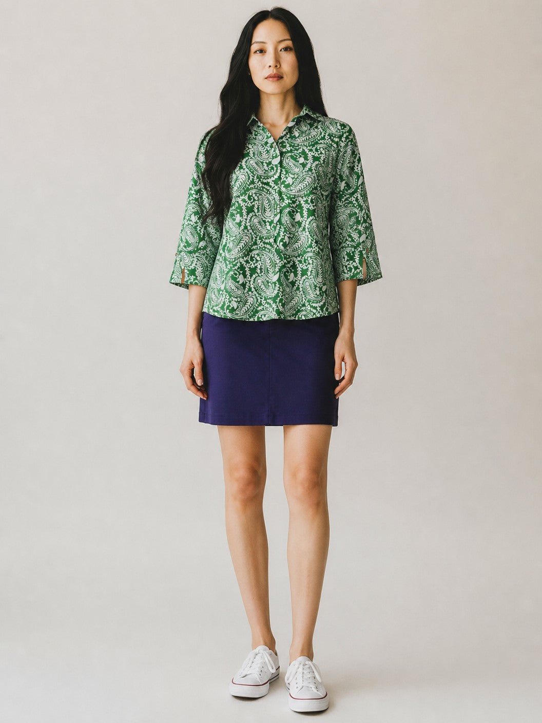 A woman modeling a green and white patterned short-sleeve button-up shirt paired with a dark navy blue mini skirt and white sneakers, standing against a plain light gray studio background.