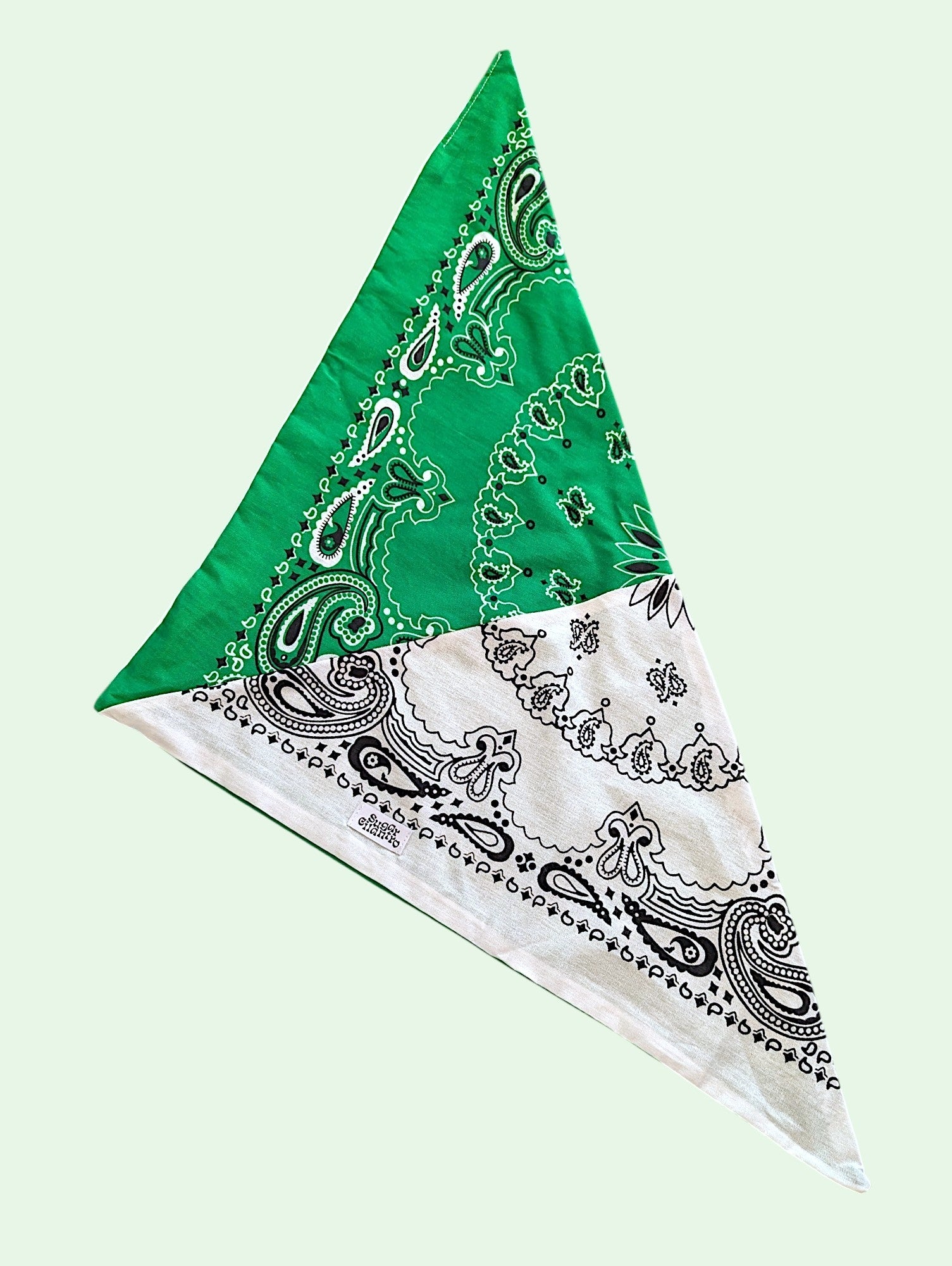 A triangular bandana featuring a split design with a bright green top section and a white bottom section, both decorated with black paisley and floral border patterns, shown on a pale mint green background.