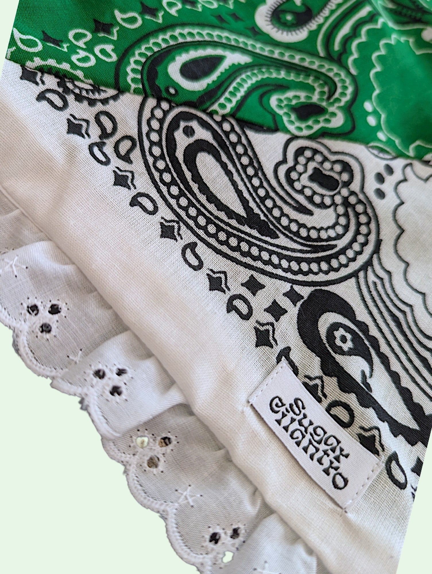A close-up of a paisley bandana scarf with a scalloped eyelet lace border. The scarf features a green, white, and black paisley pattern in the top section, and a white, black, and gray paisley pattern with matching scalloped lace along the bottom.