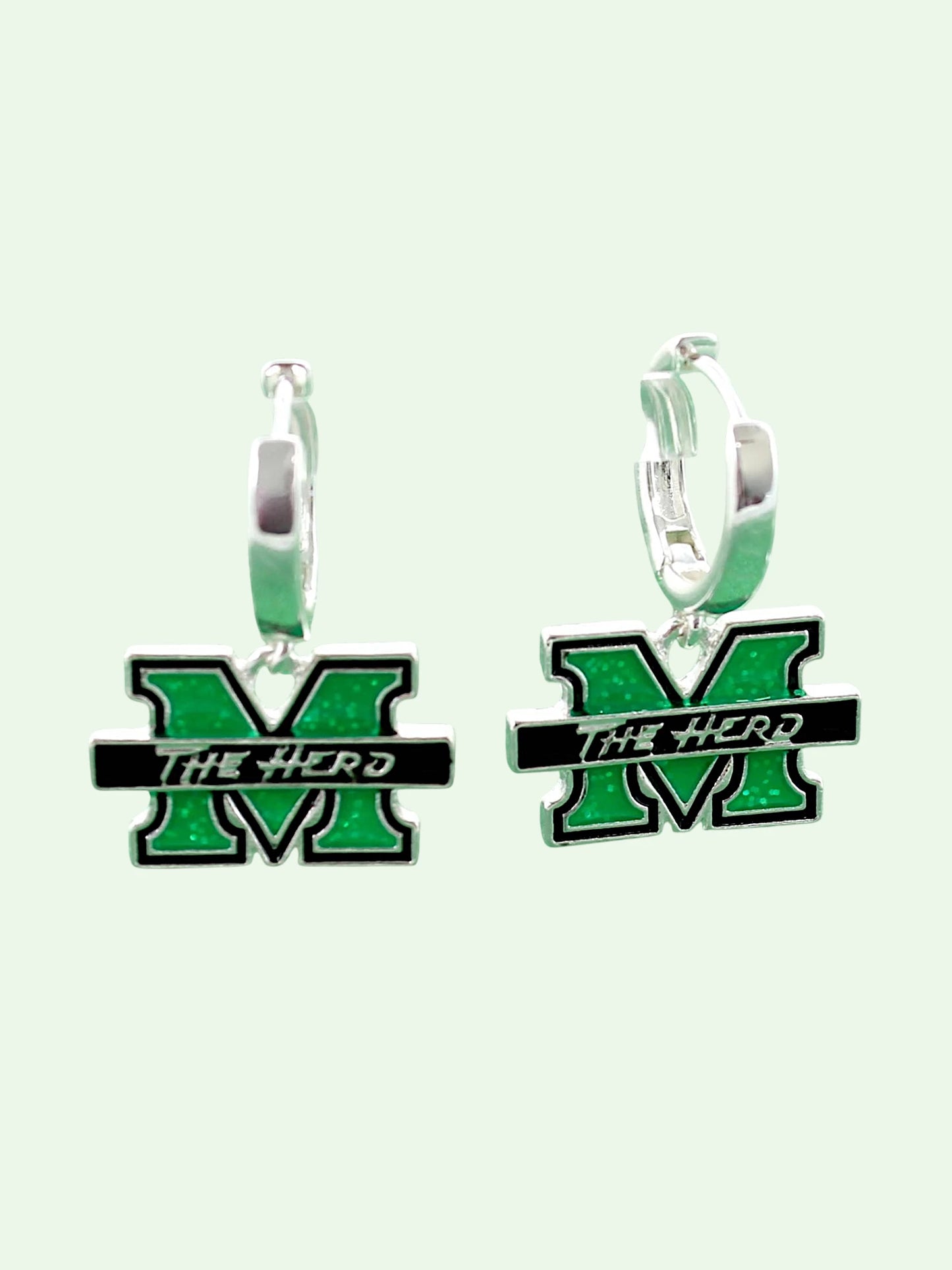 A pair of silver huggie hoop earrings featuring a green and black enamel 'M' charm with the text 'THE HERD' across a black bar, set against a pale mint green background.