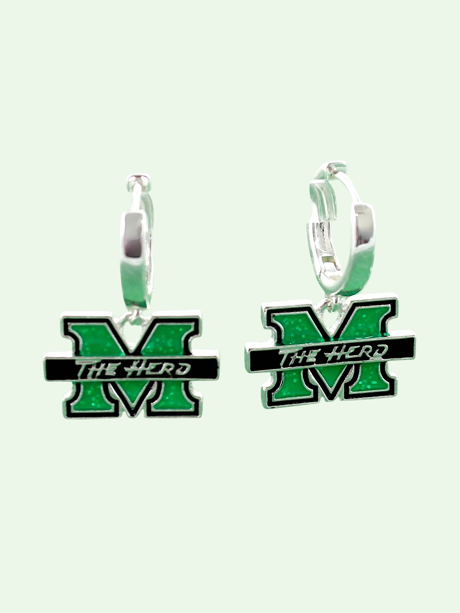 A pair of silver huggie hoop earrings featuring a green and black enamel 'M' charm with the text 'THE HERD' across a black bar, set against a pale mint green background.