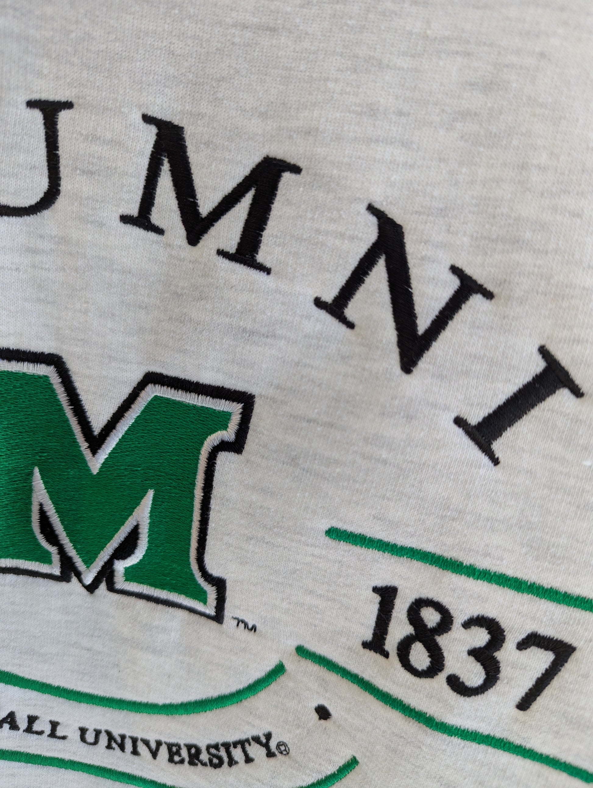 Close-up of a gray sweatshirt with 'Alumni' and '1837' text, featuring a green 'M' logo.