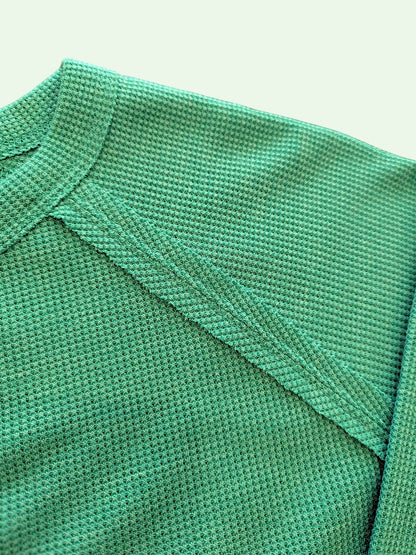 Close-up of a textured green knit fabric, showing the fine woven pattern and a diagonal seam on a light off-white background.