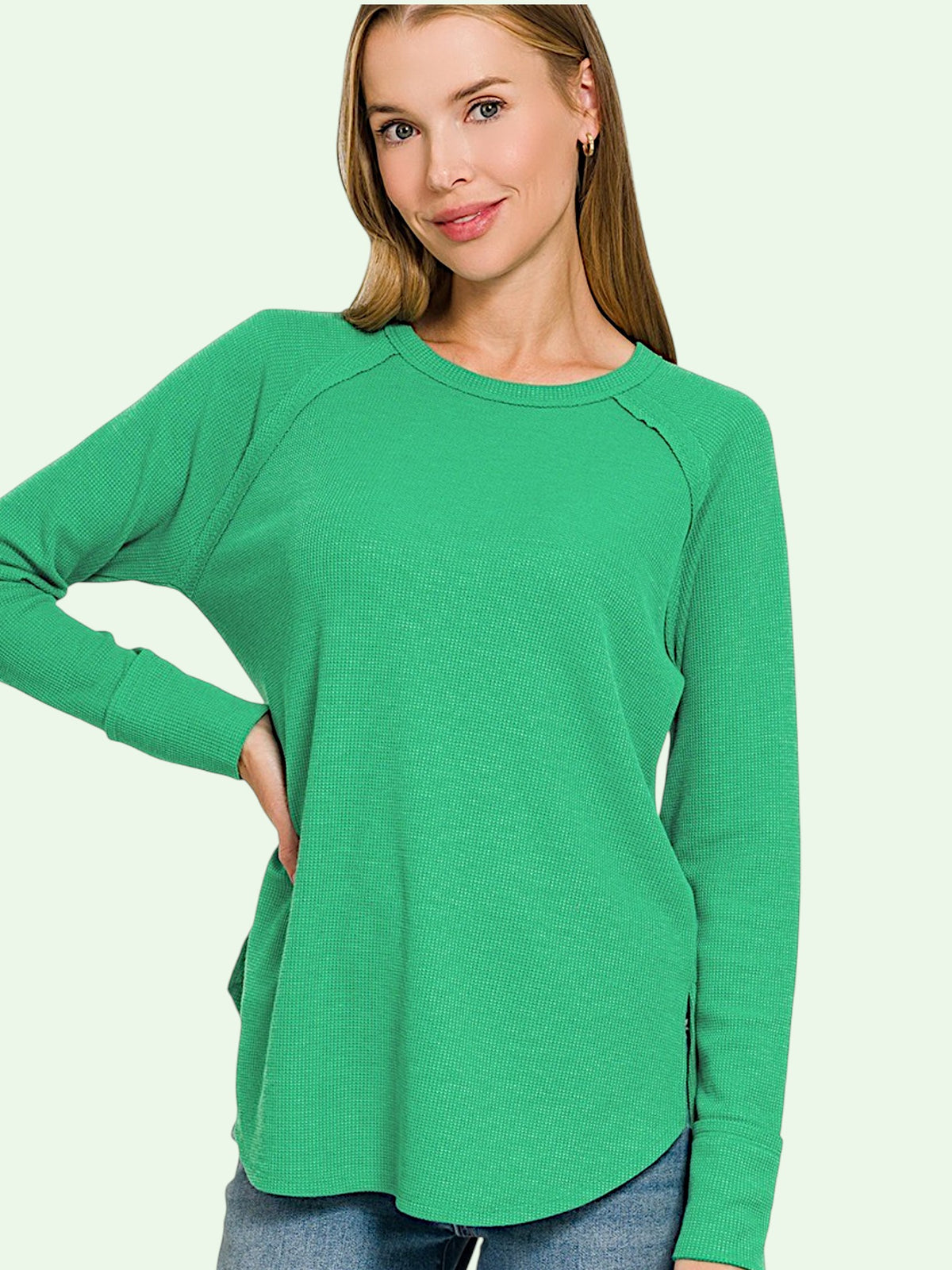 A woman wearing a bright green long-sleeve raglan top with side pockets, paired with blue jeans, against a solid pale green background.