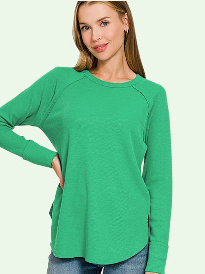 A woman wearing a bright green long-sleeve raglan top with side pockets, paired with blue jeans, against a solid pale green background.