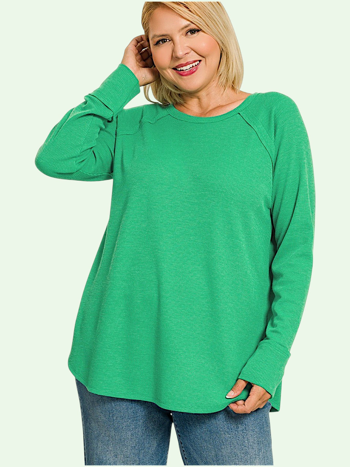 A woman wearing a bright green long-sleeve raglan top and blue jeans, posing against a light off-white background.