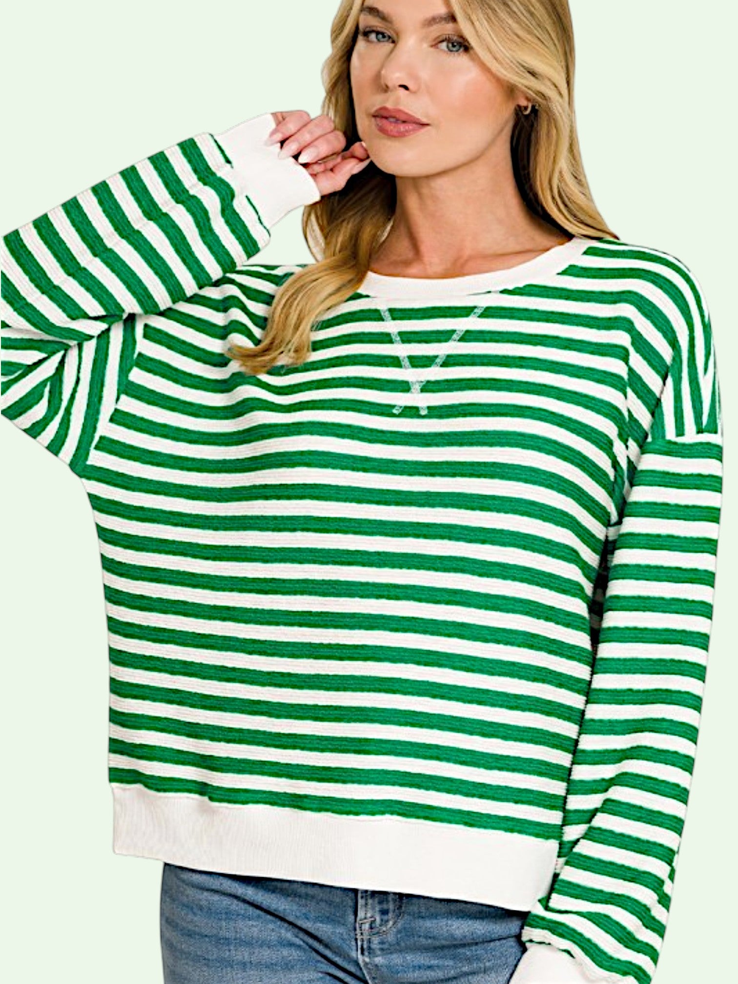 A woman wearing a green and white horizontally striped long-sleeve sweater with a crew neck and ribbed hem, paired with blue jeans against a pale green background.