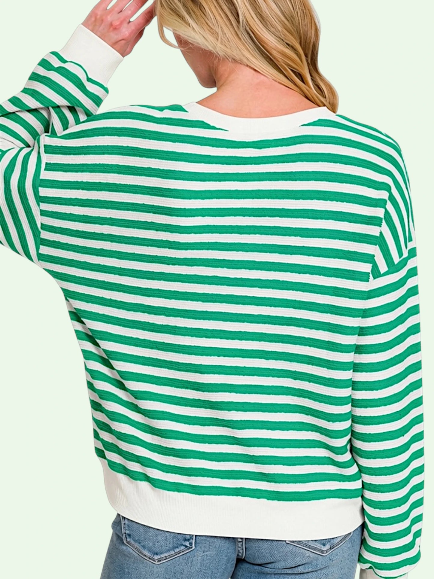 A woman shown from the back wearing a green and white horizontally striped long-sleeve sweater with solid white ribbed cuffs, collar, and hem, paired with blue jeans against a pale mint green background.