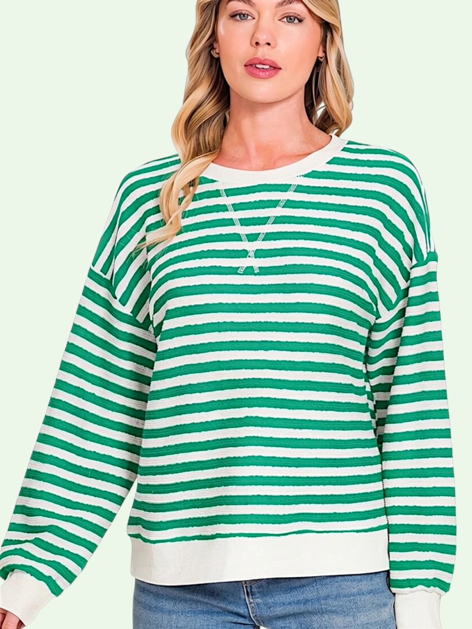 A woman wearing a green and white horizontally striped long-sleeve crewneck sweater, paired with blue jeans, against a solid pale green background.