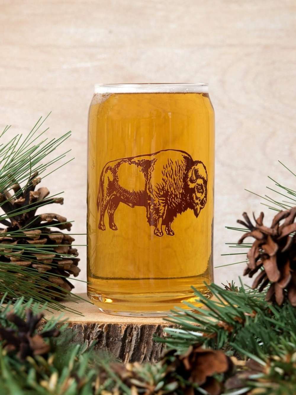 Glass with bison design filled with amber liquid, surrounded by pine branches and pinecones on a wooden surface.