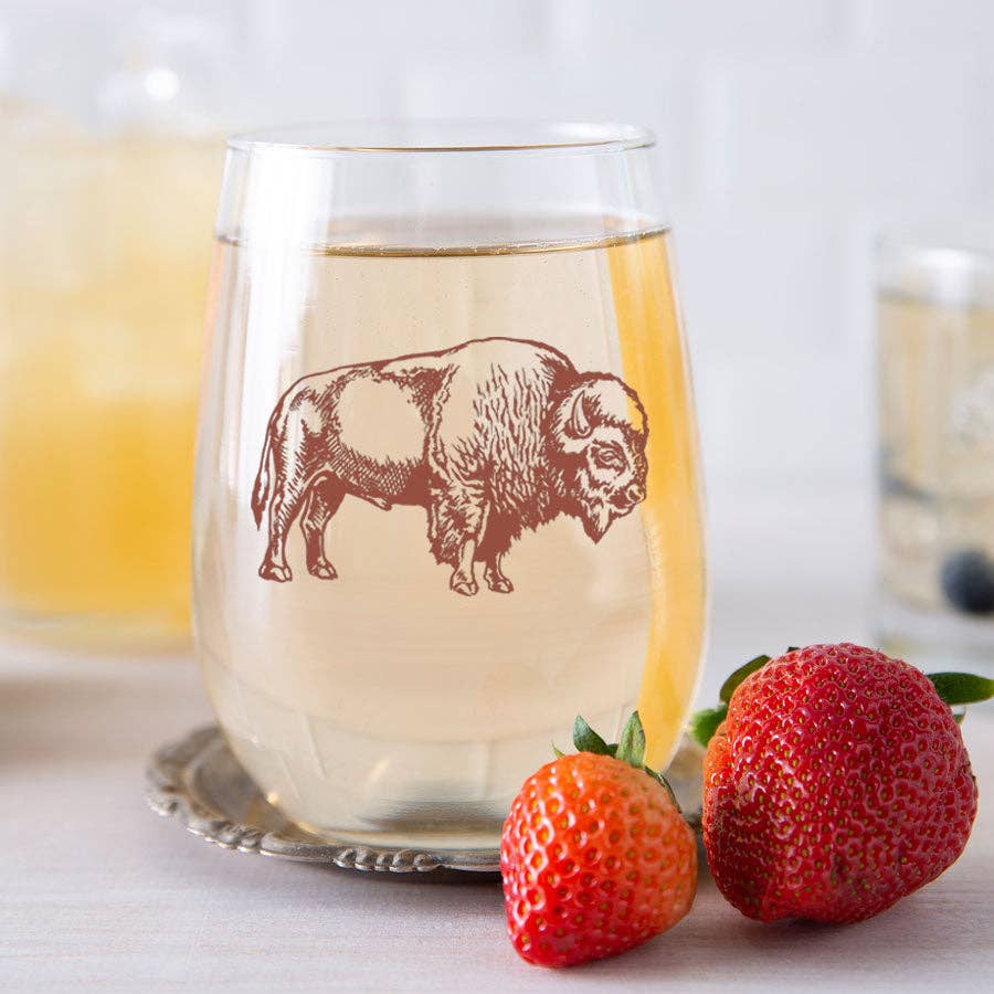 Glass of white wine with a bison illustration, surrounded by strawberries on a white surface.