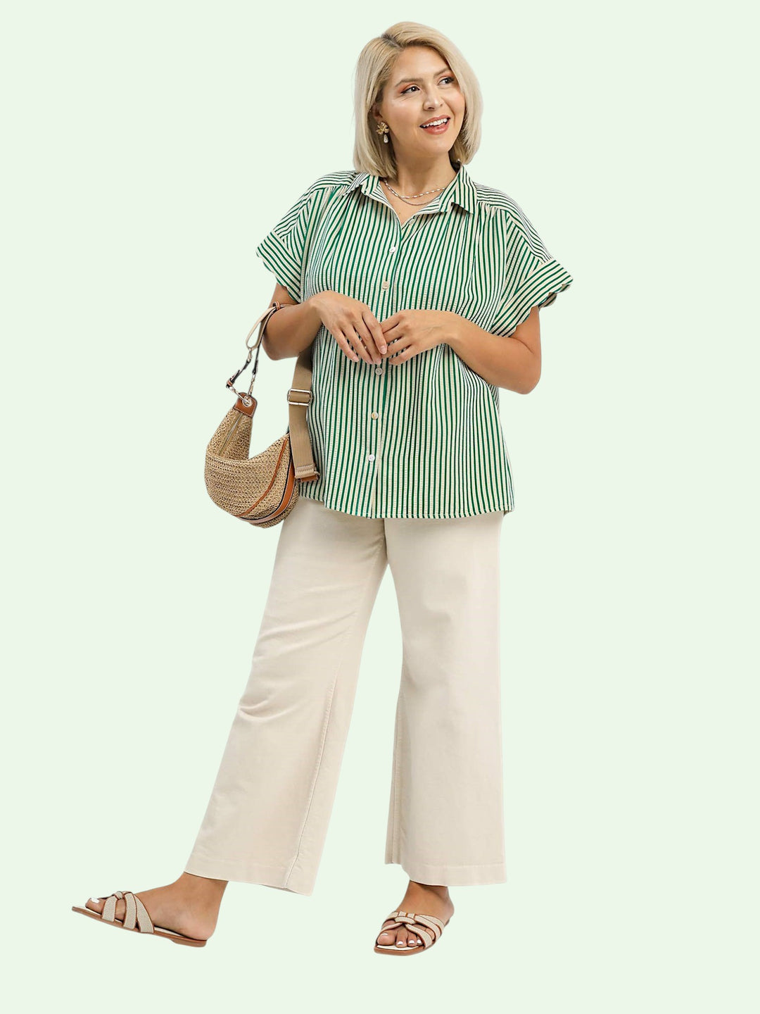 Woman wearing a green and white striped shirt and beige pants, holding a brown bag on a light background