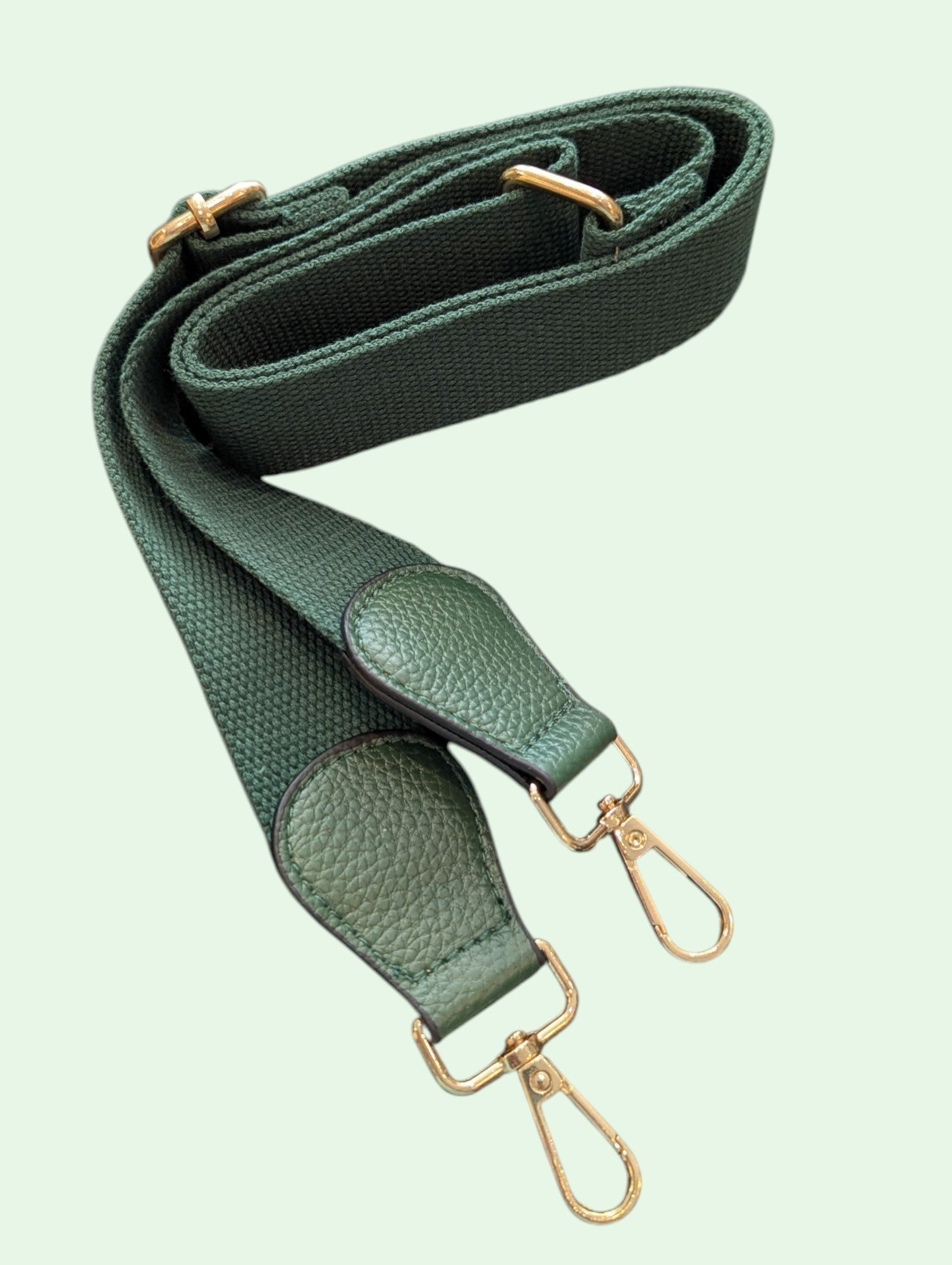 A dark green textured fabric shoulder strap with gold-tone metal hardware, including swivel clips and adjustment loops, displayed on a pale mint green background.