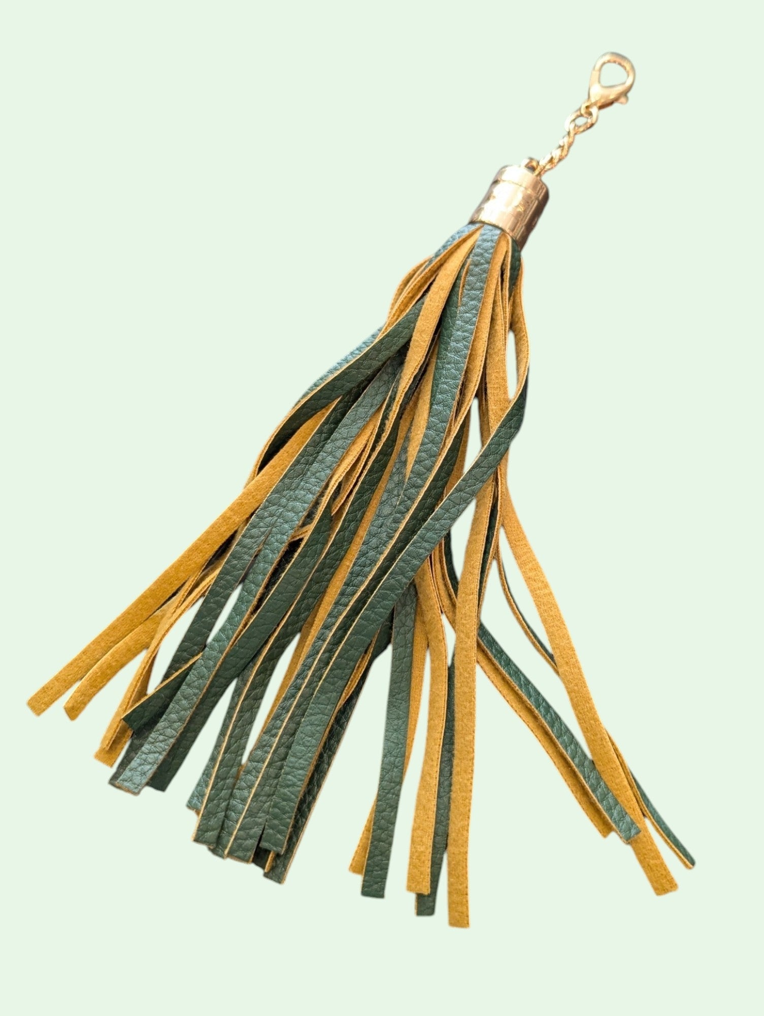 A leather tassel keychain featuring alternating green and mustard yellow fringe strips, finished with a gold-tone metal cap and lobster clasp, set against a pale mint green background.