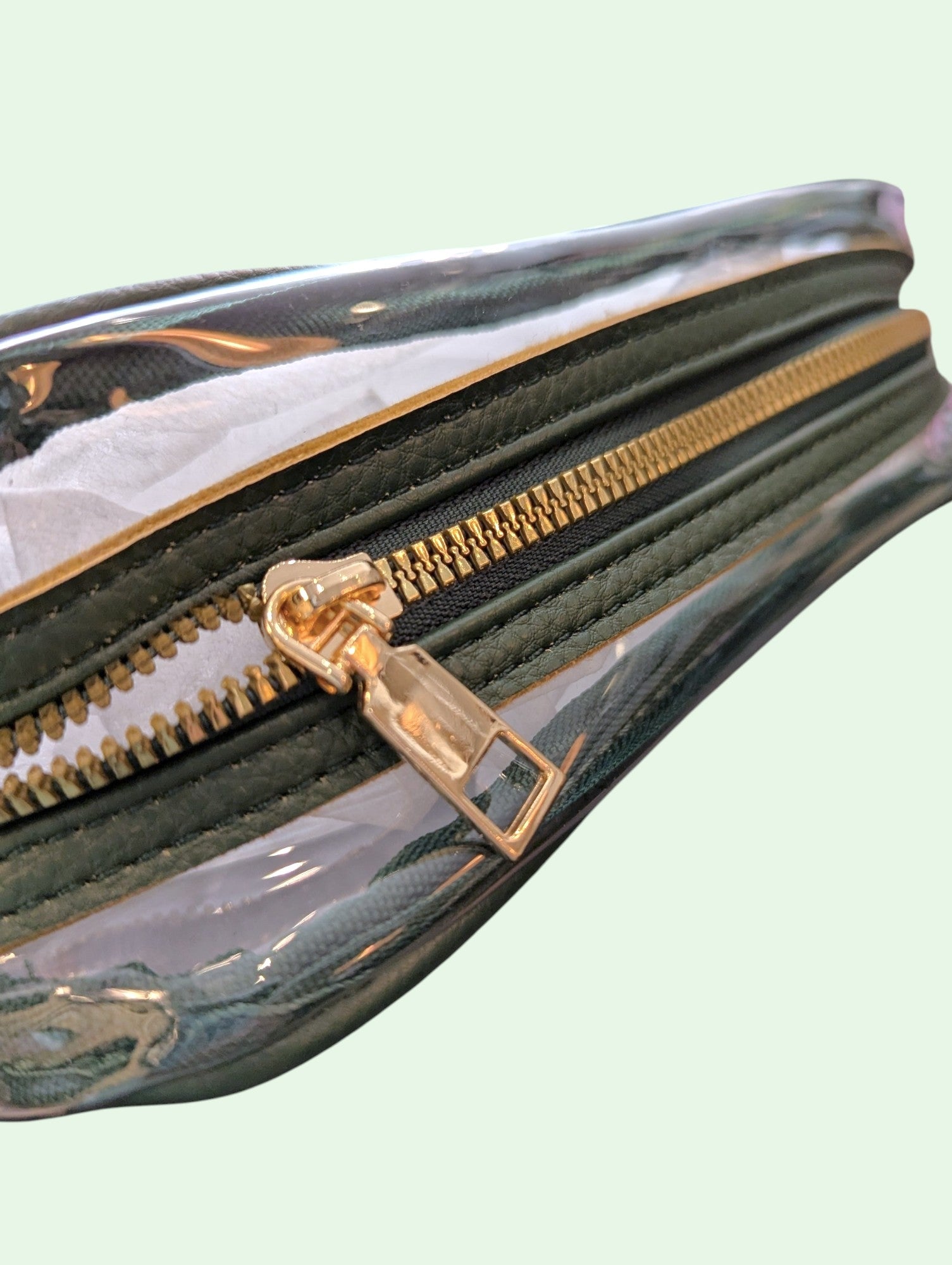 A close-up of a clear vinyl pouch with a gold zipper and a dark olive green fabric lining, shown against a pale mint green background.