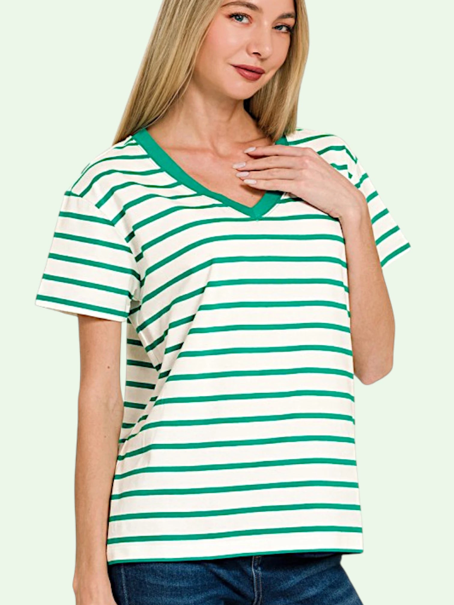 A woman wearing a green and white horizontally striped short-sleeve V-neck t-shirt with solid green trim, paired with blue jeans, against a pale mint green background.