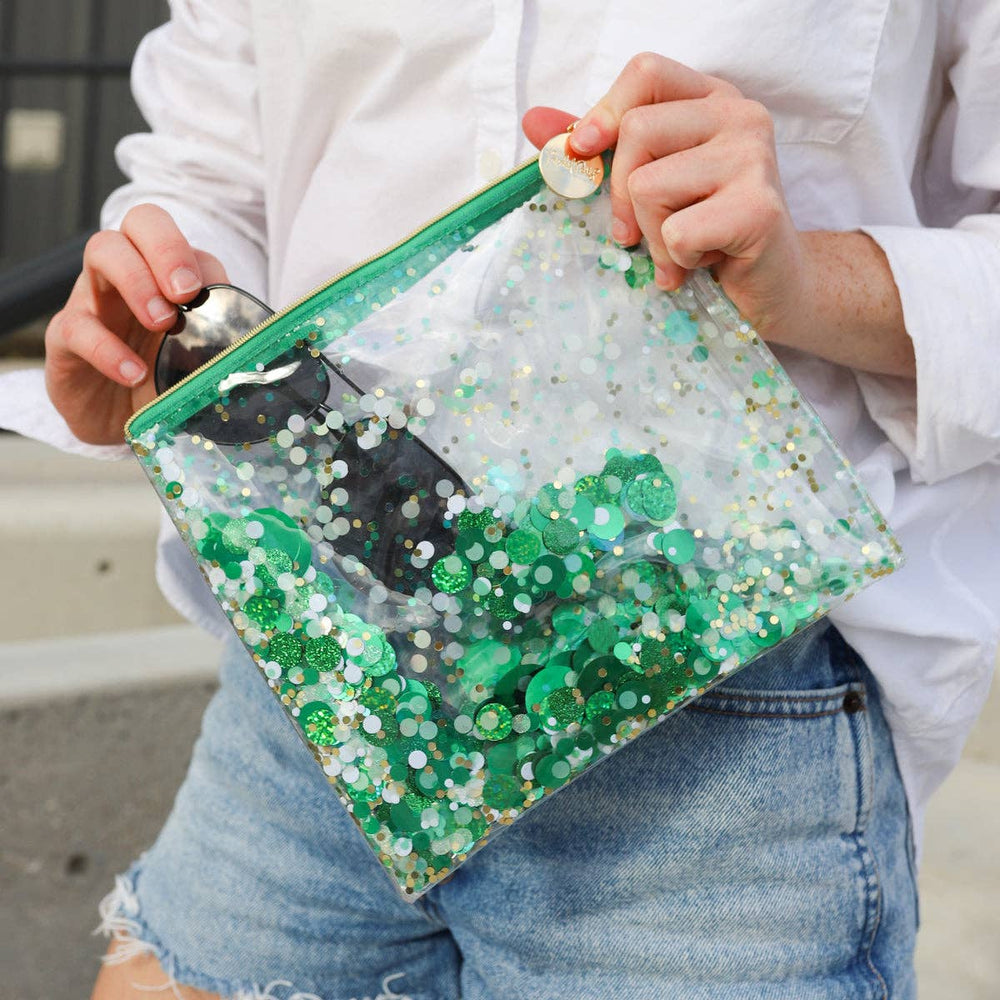 Person holding a clear pouch with green glitter and a camera inside, wearing a white shirt and blue jeans.