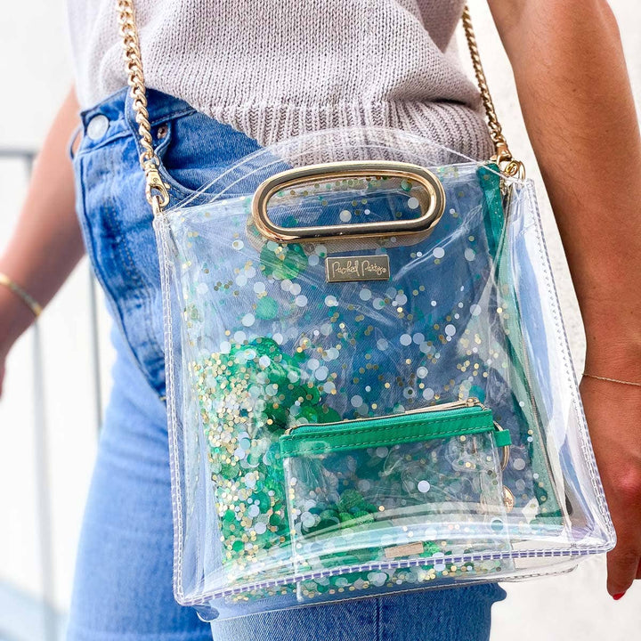 Clear handbag with green glittery interior, gold clasp, and chain strap worn by a person.