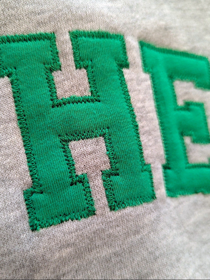 Close-up of a light heather gray fabric with large green embroidered letters 'HE' shown in detail.