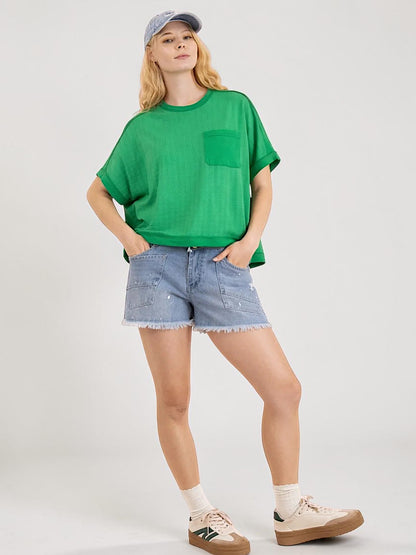 A woman modeling a green short-sleeve crewneck t-shirt with a chest pocket, paired with light-wash denim shorts, white socks, and beige sneakers, set against a plain light gray studio background.