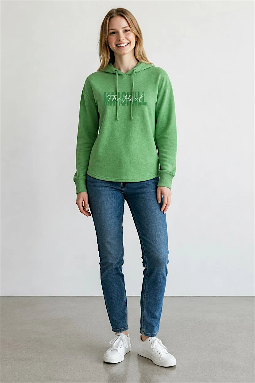 A woman modeling a green long-sleeve hoodie with a white graphic text print on the chest, paired with blue jeans and white sneakers, standing against a plain light gray wall.