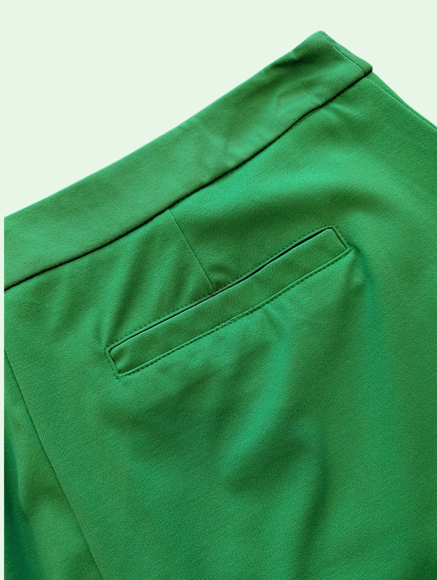 Close-up of bright green trousers showing the waistband, a side pocket, and fabric texture against a pale mint background.