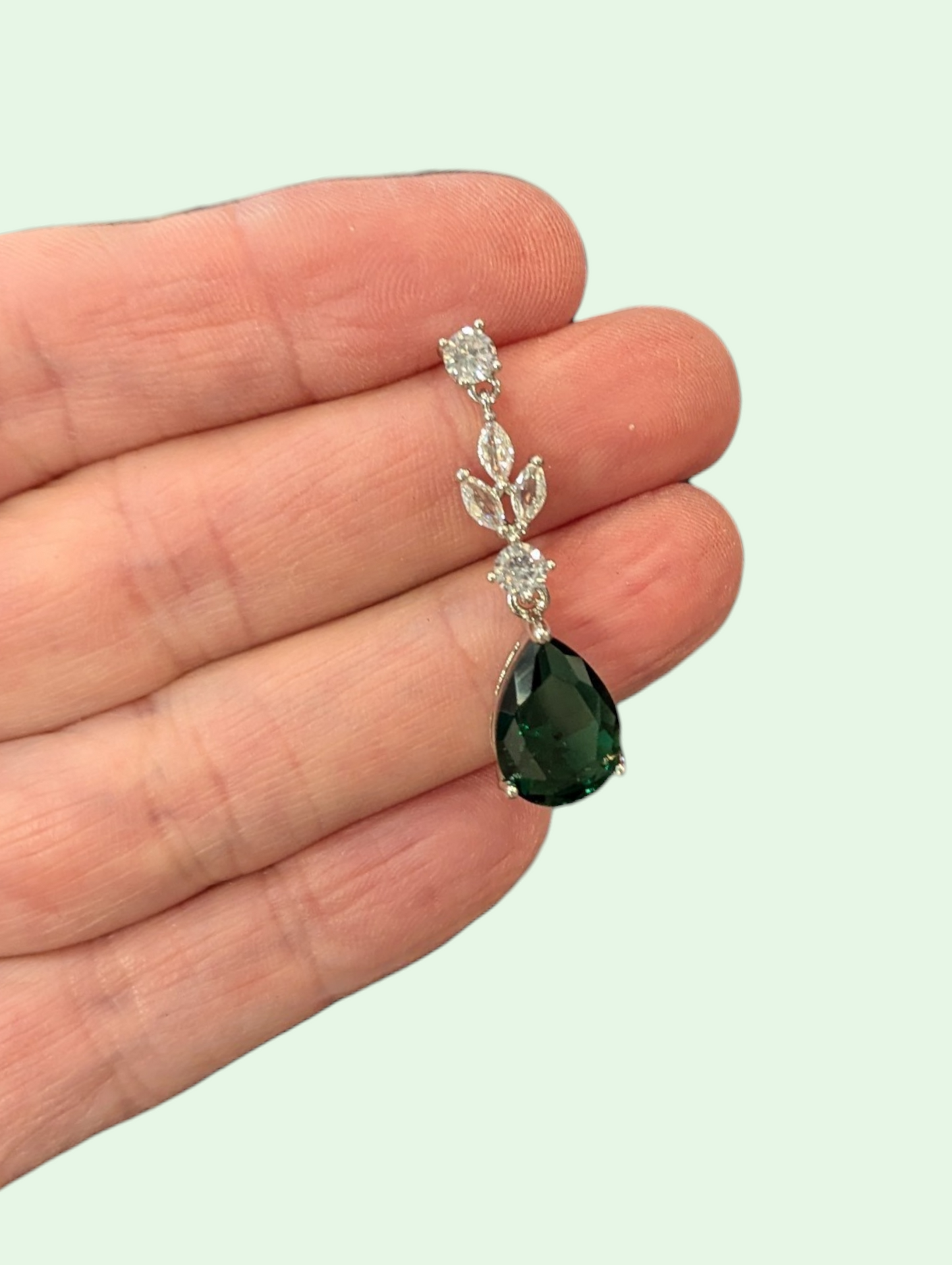 Green teardrop earring with silver accents held in a hand against a light background