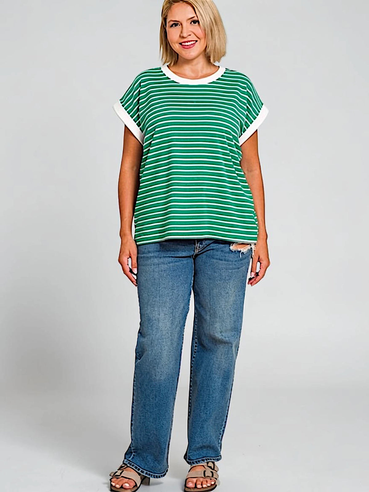 A woman modeling a green and white horizontally striped short-sleeve t-shirt with white ribbed trim on the collar and sleeve cuffs, paired with blue jeans and brown sandals, against a light gray studio background.