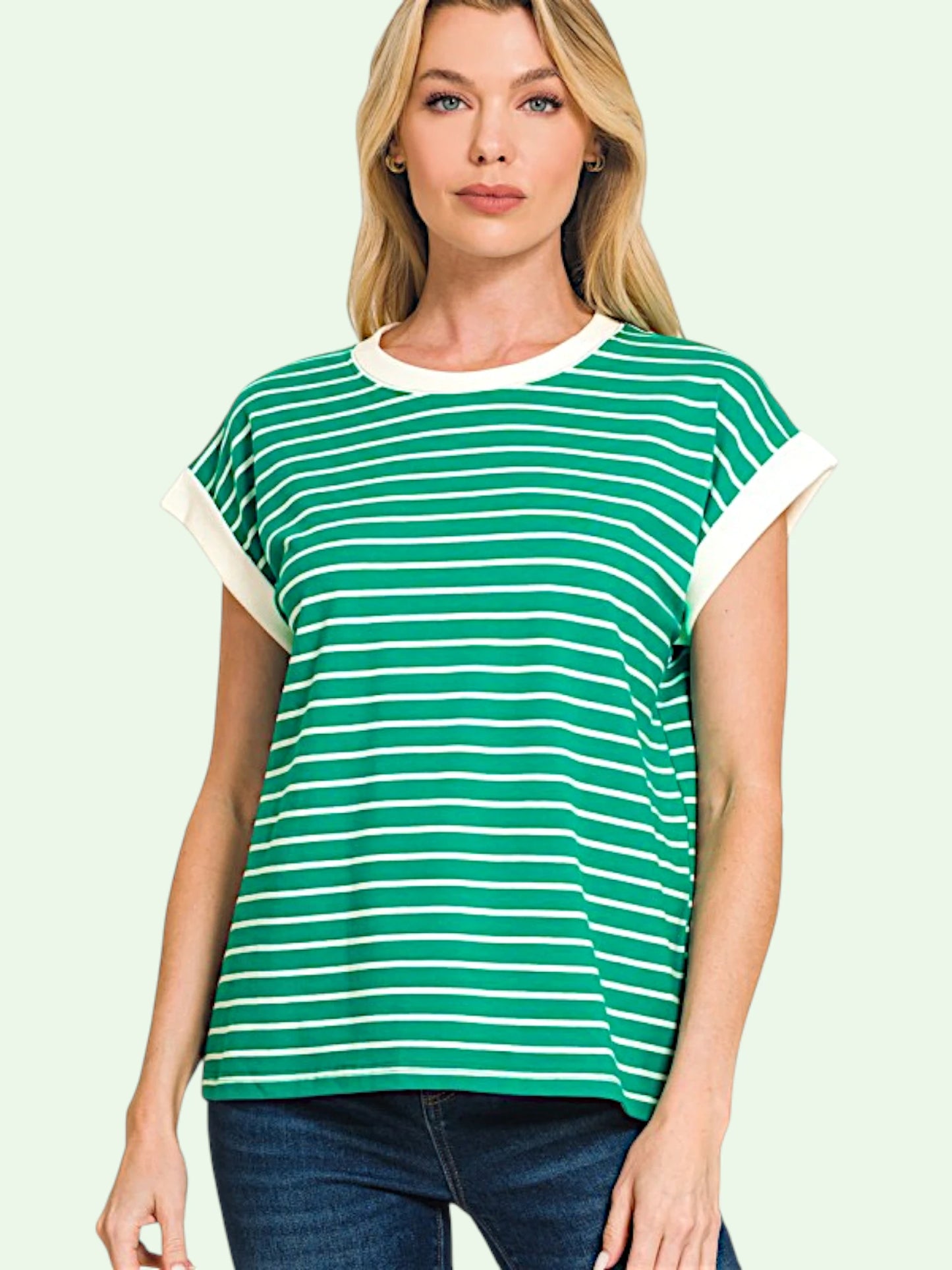 A woman wearing a green and white horizontally striped short-sleeve t-shirt with solid white ribbed trim at the crew neck and sleeve cuffs, paired with dark blue jeans, set against a solid pale green background.
