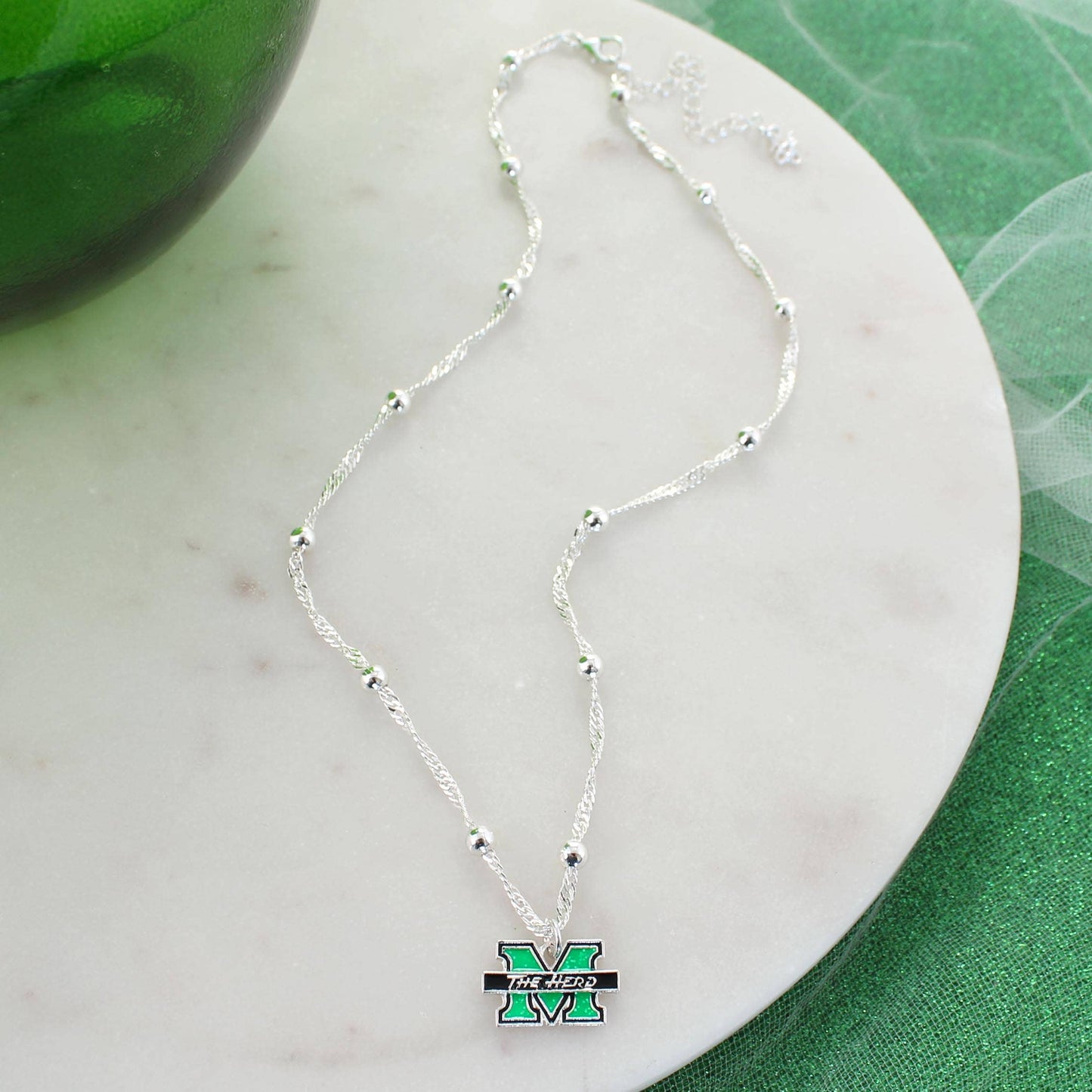 A silver beaded chain necklace with a green and black Michigan 'M' logo pendant, displayed on a white marble coaster over a green textured surface.
