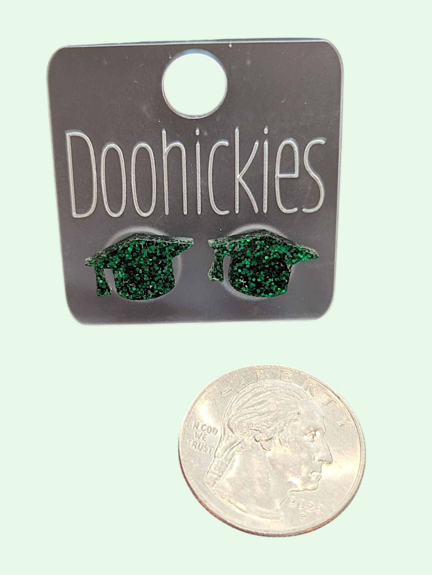 Green graduation cap earrings on a 'Doohickies' card with a quarter for scale.
