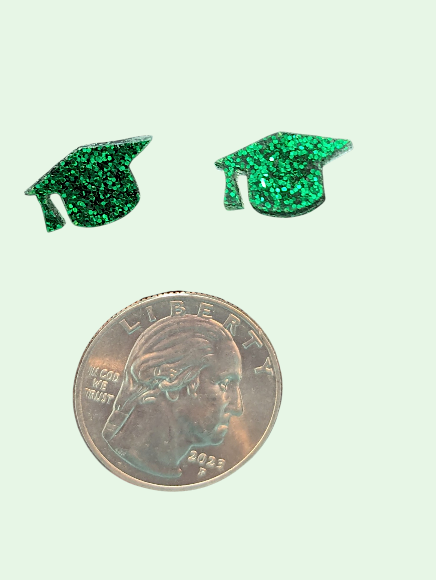 Green glittery graduation cap earrings next to a quarter on a light green background