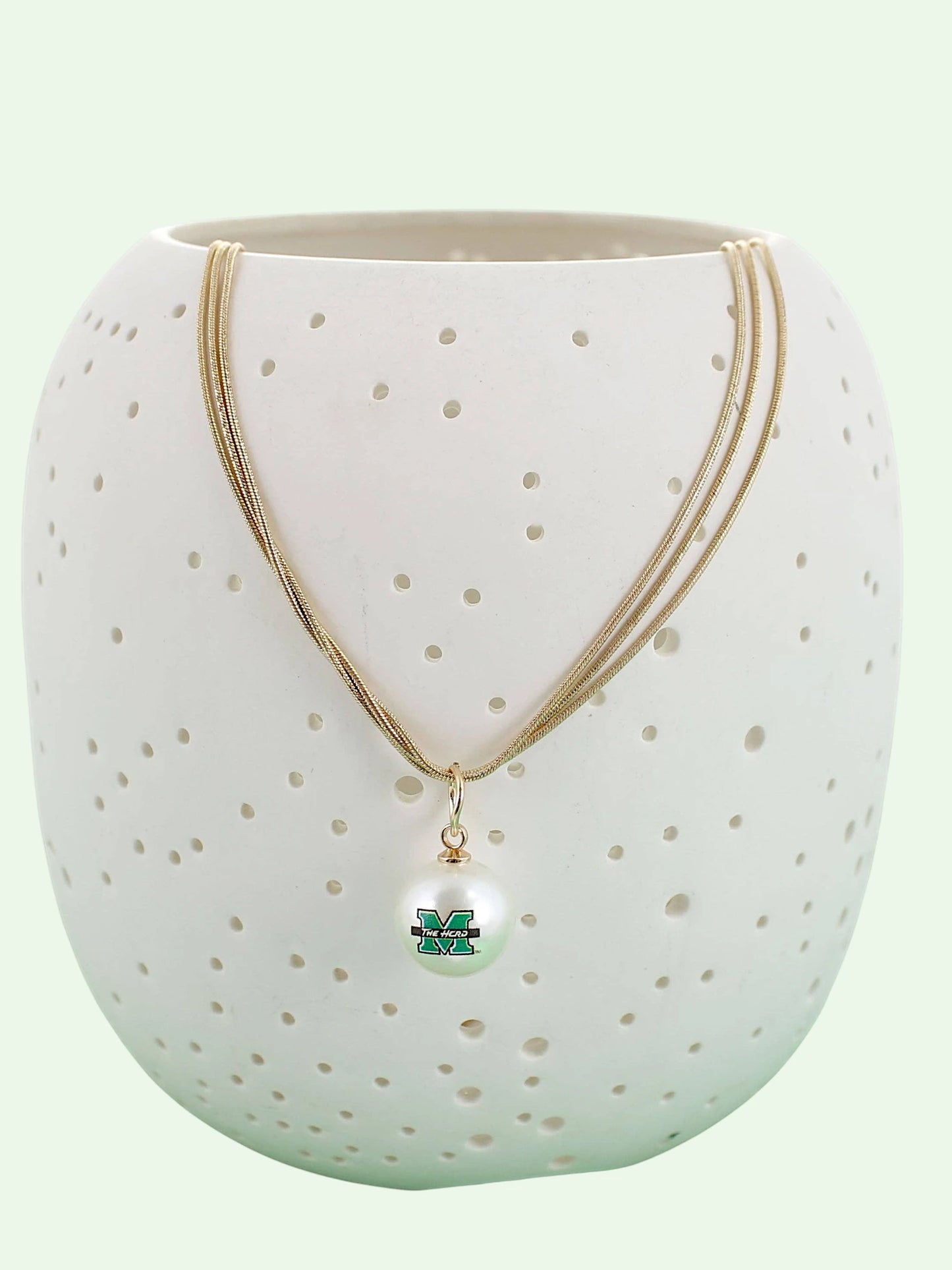 A gold multi-strand necklace with a white pearl pendant featuring a green and Marshall 'M' logo, displayed on a white ceramic vase with perforated holes against a pale mint green background.