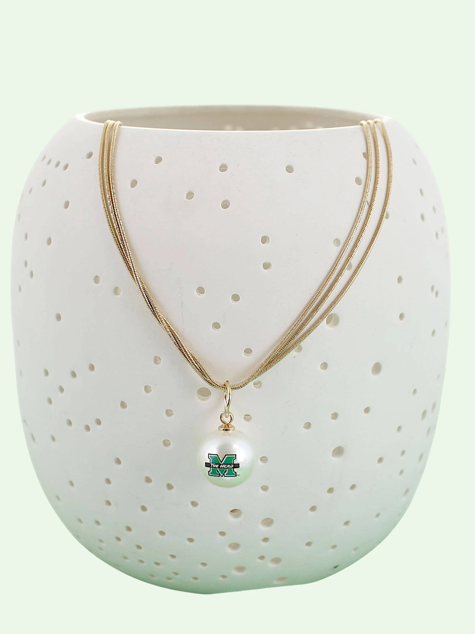 A gold multi-strand necklace with a white pearl pendant featuring a green and Marshall 'M' logo, displayed on a white ceramic vase with perforated holes against a pale mint green background.
