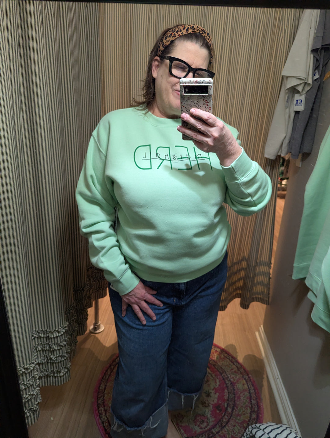 Person wearing a light green sweatshirt with text and blue jeans, taking a mirror selfie.
