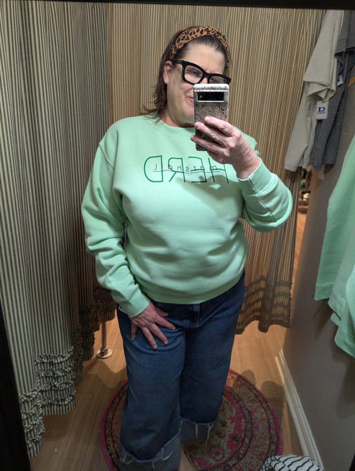 Person wearing a light green sweatshirt with text and blue jeans, taking a mirror selfie.