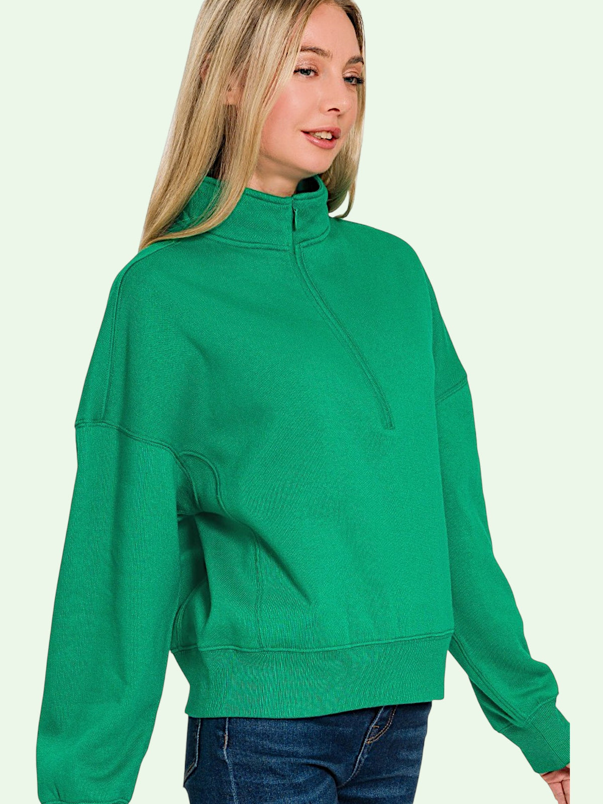 A woman wearing a bright green mock-neck long-sleeve sweatshirt with a half-zip front, paired with blue jeans, set against a solid pale green background.
