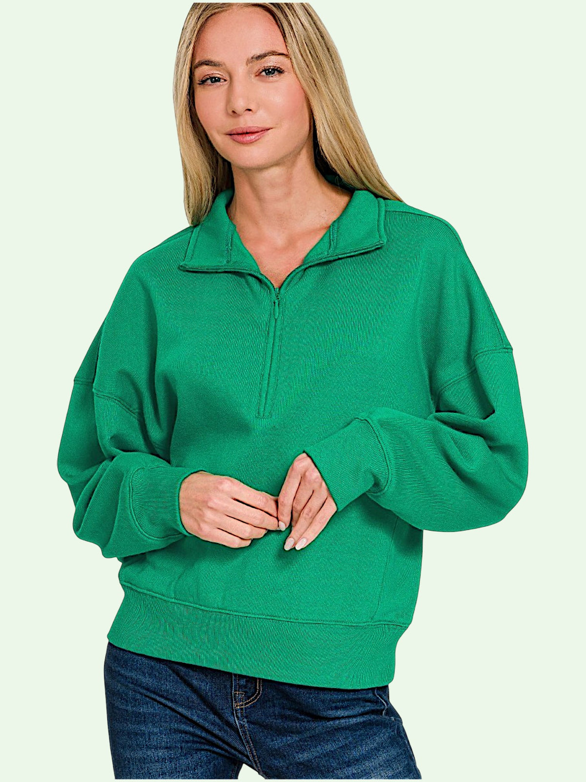 A woman wearing a bright green half-zip sweatshirt with a mock neck and long sleeves, paired with blue jeans, against a pale mint green background.