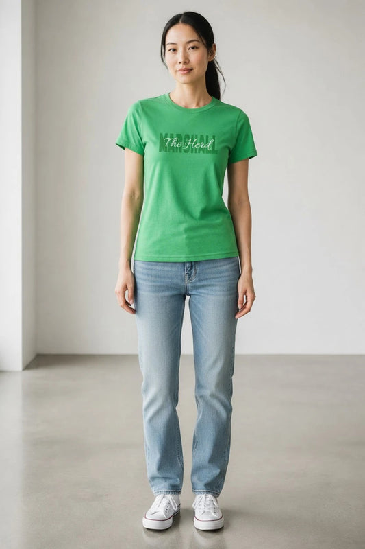 A woman stands in a minimalist studio wearing a bright green short-sleeve t-shirt with a dark green graphic text on the chest, paired with light-wash blue jeans and white sneakers.