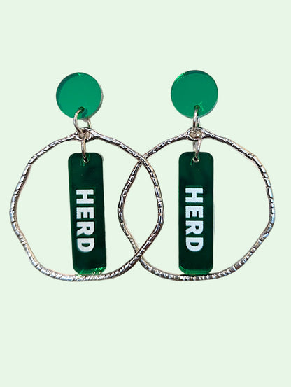 A pair of statement dangle earrings featuring textured silver-tone wavy hoop frames, a round green disc, and a vertical green bar with the word 'HERD' printed in white, set against a pale mint green background.
