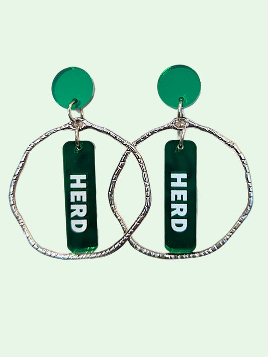 A pair of statement dangle earrings featuring textured silver-tone wavy hoop frames, a round green disc, and a vertical green bar with the word 'HERD' printed in white, set against a pale mint green background.