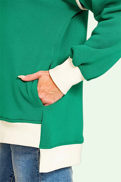 A close-up of a person wearing a green long-sleeve top with contrasting cream ribbed cuffs and hem, with a hand tucked into a side pocket, paired with blue jeans against a pale mint background.