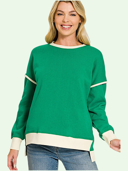 A smiling blonde woman wearing a green crewneck sweatshirt with cream ribbed trim on the collar, cuffs, hem, and sleeve details, paired with blue jeans against a pale mint green background.