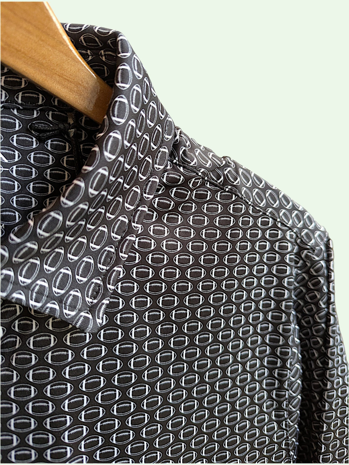 A close-up of a black football patterned shirt with a white geometric repeating pattern, hanging on a wooden hanger against a pale mint green background.