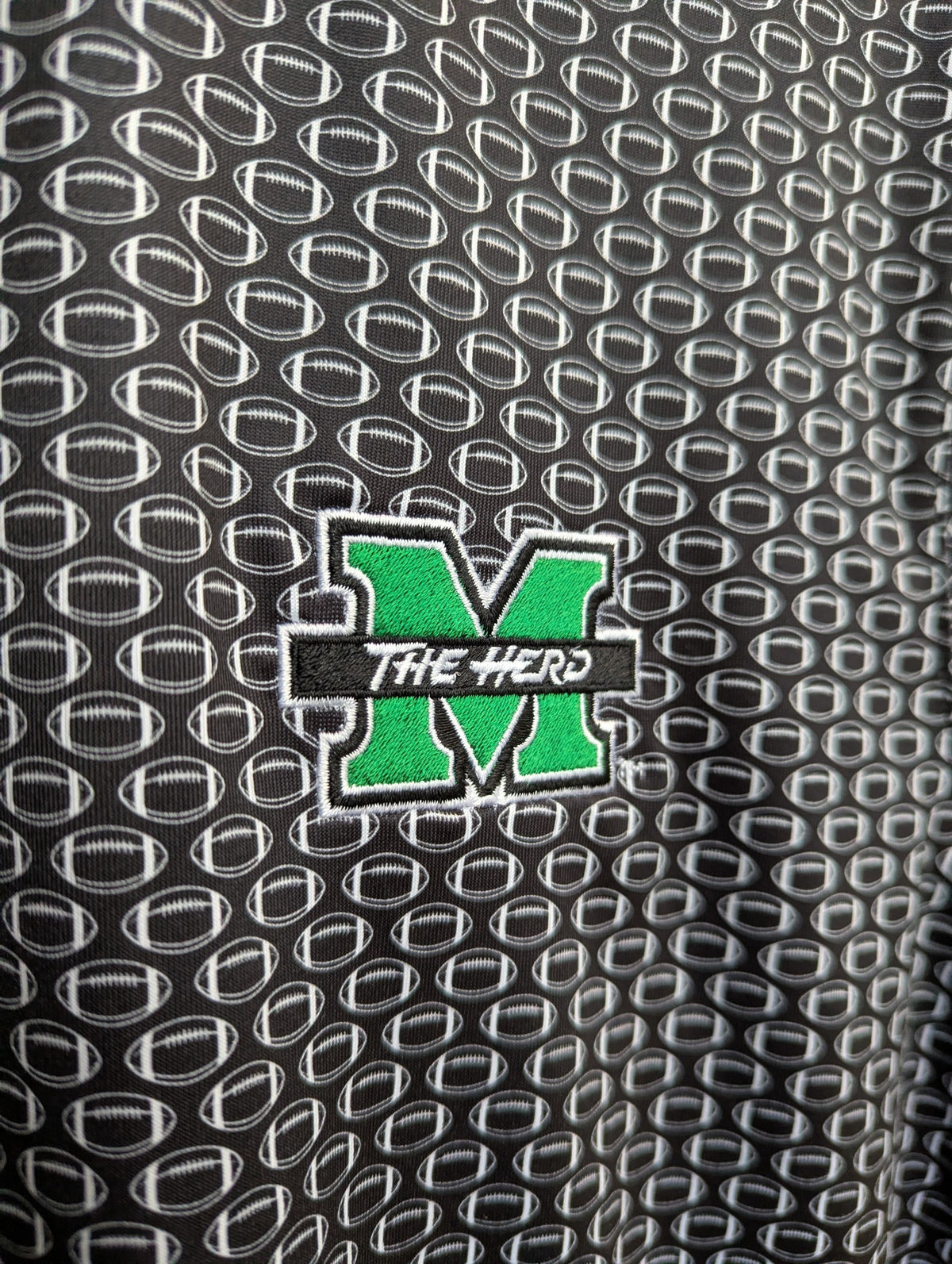Close-up of a black  polo with a repeating white football pattern and a green embroidered letter M with the words 'THE HERD' across the center.