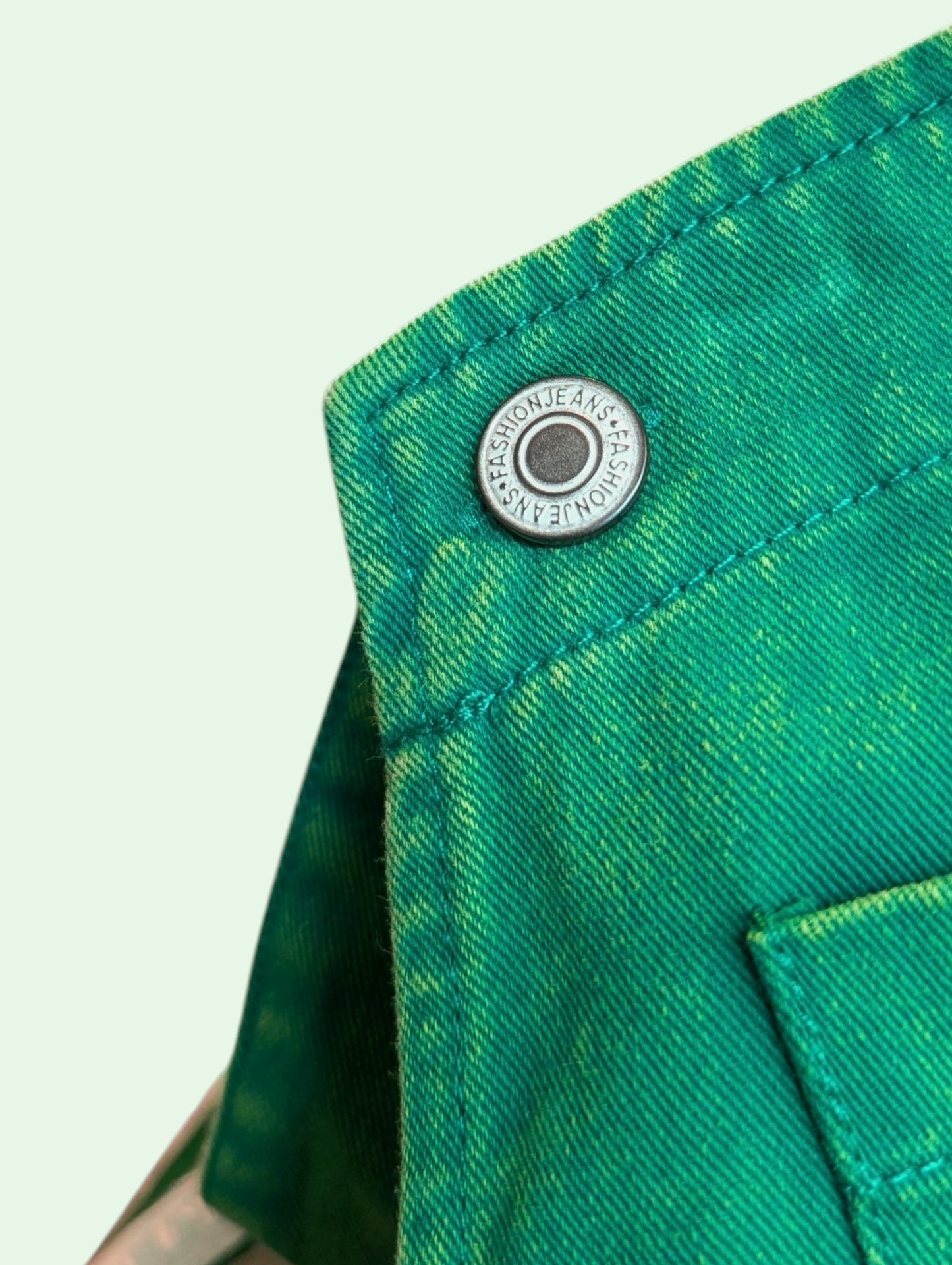 Close-up of a bright green denim garment showing a silver metal snap button with the brand name 'FASHION JEANS' printed on it, set against a pale mint green background.