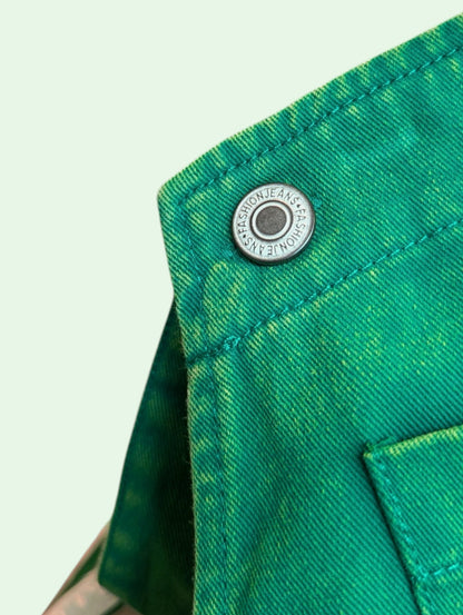 Close-up of a bright green denim garment showing a silver metal snap button with the brand name 'FASHION JEANS' printed on it, set against a pale mint green background.