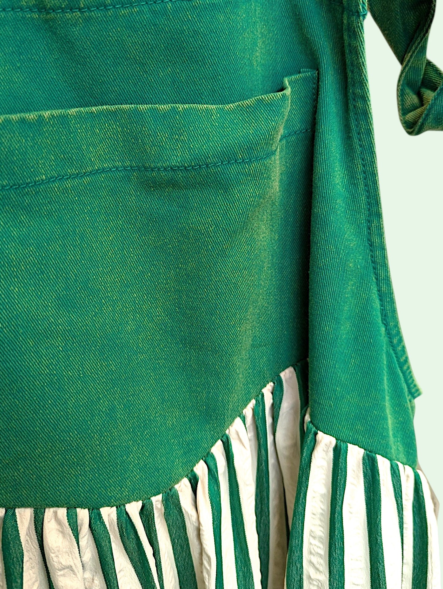 Close-up of a green denim garment, showing a pocket on the front and a skirt with alternating green and off-white.